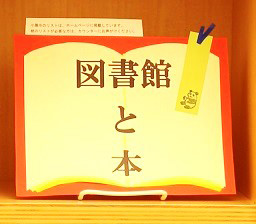 小展示「図書館と本」の看板の写真。開かれた本を模したイラストが描かれ、その中に展示タイトル「図書館と本」が印字されている。看板の右上には栞が貼り付けられている。